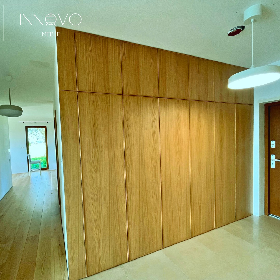 Jasna, drewniana szafa wnękowa od podłogi do sufitu, zlicowana ze ścianą, w przedpokoju z drewnianą podłogą i widokiem na zewnątrz.