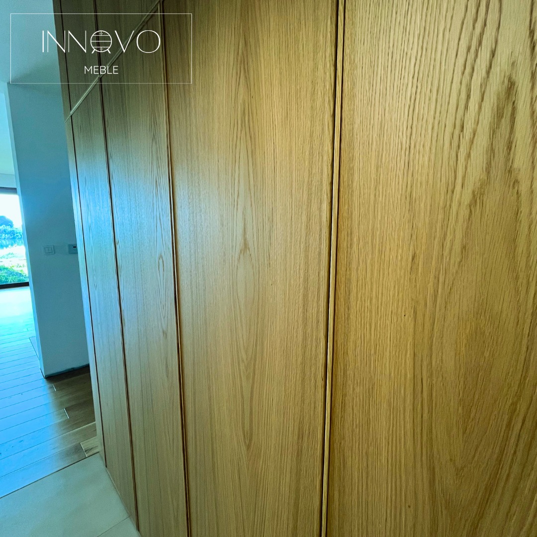 Jasna, drewniana szafa wnękowa z subtelnymi, pionowymi podziałami, dopasowana do wnętrza. Widoczna część korytarza z oknem i zielenią w tle.