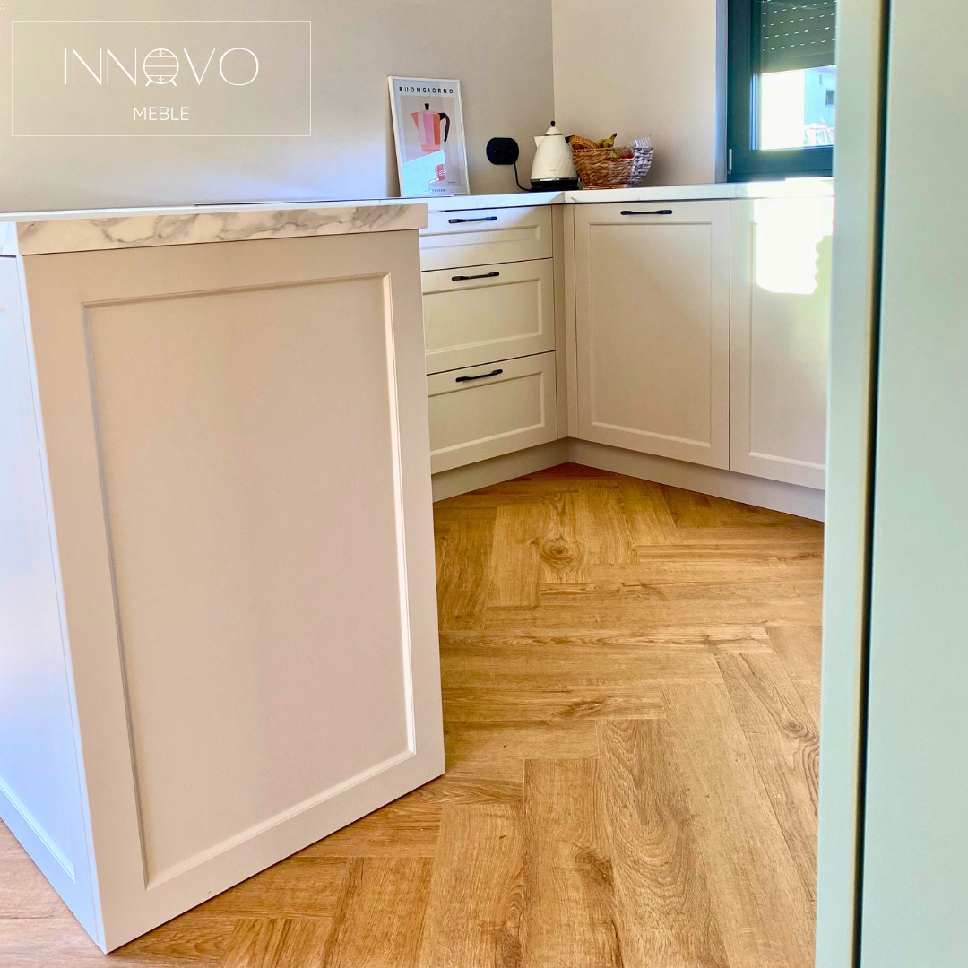 Jasna, narożna kuchnia z frezowanymi frontami i marmurowym blatem, widok na parkiet ułożony w jodełkę, czarne uchwyty, minimalistyczny plakat na blacie.