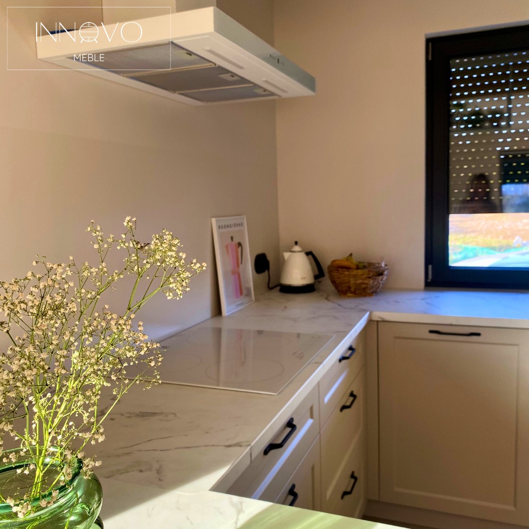 Jasna kuchnia z marmurowym blatem i kwiatami w wazonie. Widoczny okap i okno z roletami. Minimalistyczny design i ciepłe oświetlenie.