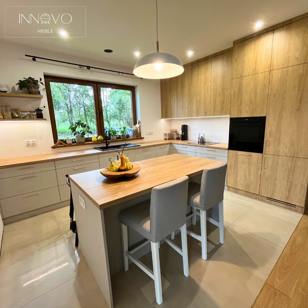 Jasna kuchnia z drewnianymi frontami i wyspą z blatem z naturalnego drewna. Na wyspie dekoracja z dyń. Nad wyspą lampa. Duże okno z widokiem na zieleń.