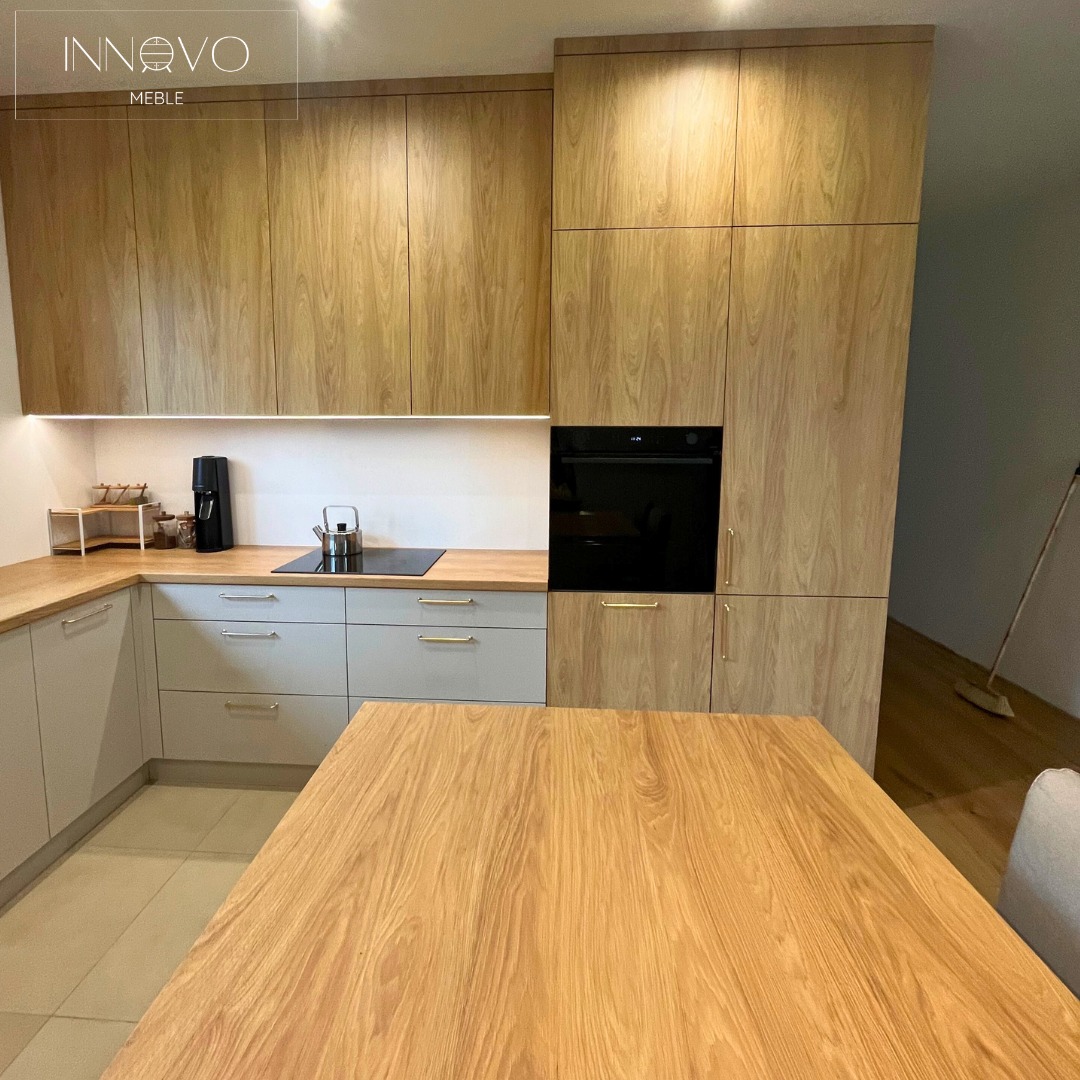 Jasna kuchnia z drewnianymi frontami i szarymi szafkami. Na blacie czajnik i ekspres do kawy. Widoczny stół z blatem o podobnym odcieniu drewna.