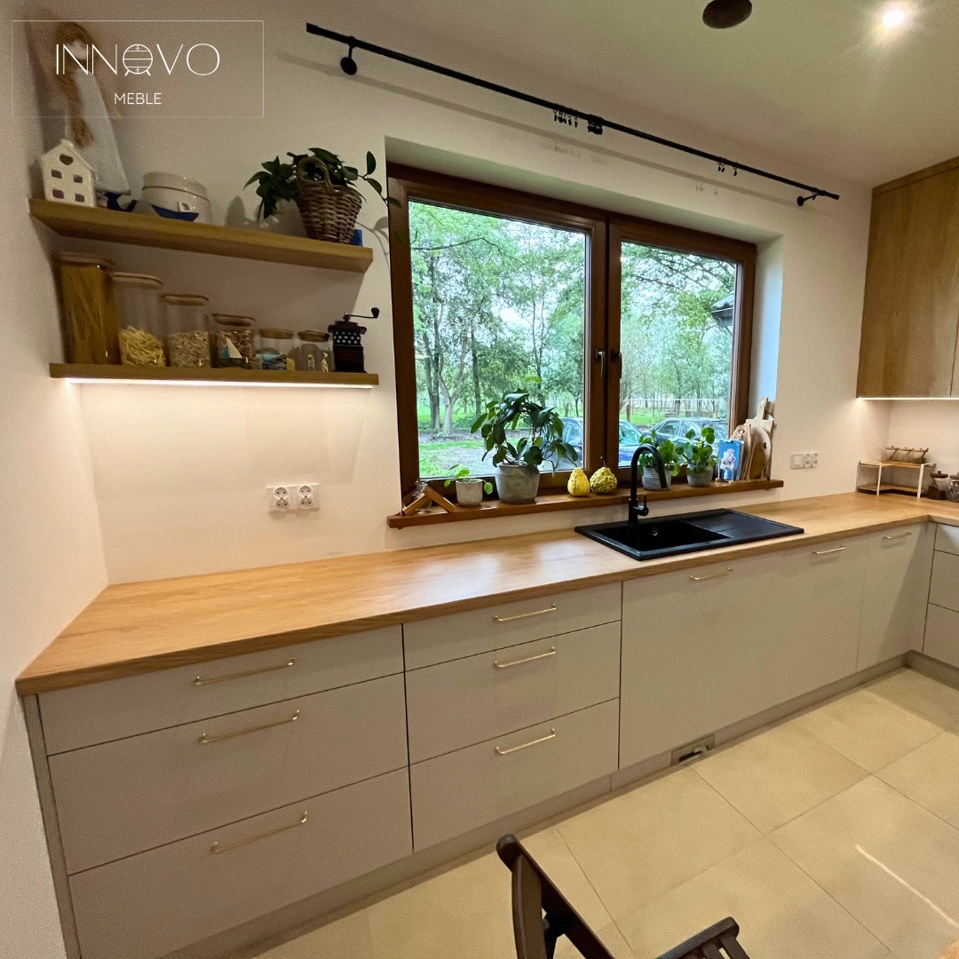 Jasna kuchnia z drewnianym blatem w Krzętowie. Szafki dolne z długimi, złotymi uchwytami. Nad blatem półki i okno z widokiem na zieleń.