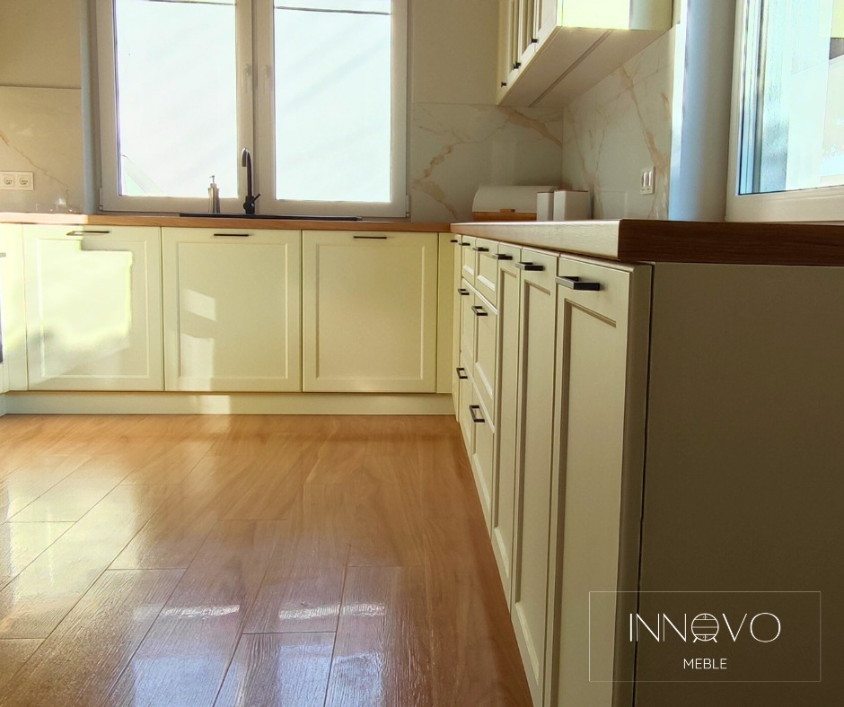 Jasna kuchnia z drewnianym blatem i frontami w stylu angielskim, czarne, podłużne uchwyty. Parkiet na podłodze. Widok z perspektywy podłogi, logo producenta w prawym dolnym rogu.