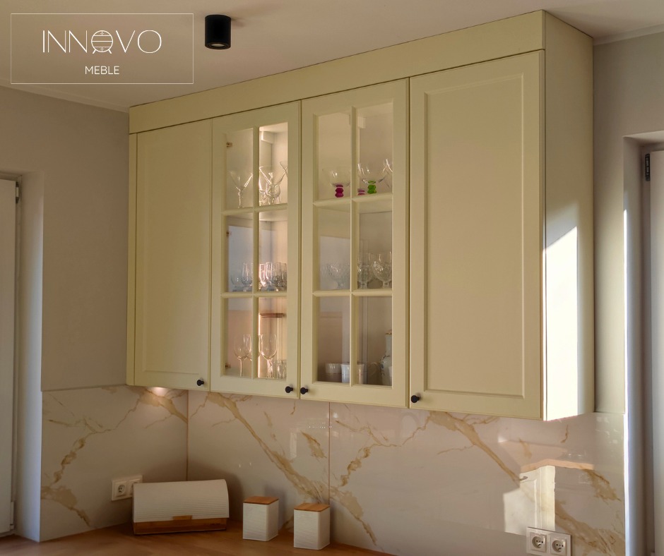 Beżowe szafki kuchenne z przeszklonymi frontami, eleganckie naczynia wewnątrz, marmurowe płytki na ścianie i drewniany blat. Minimalistyczny design.