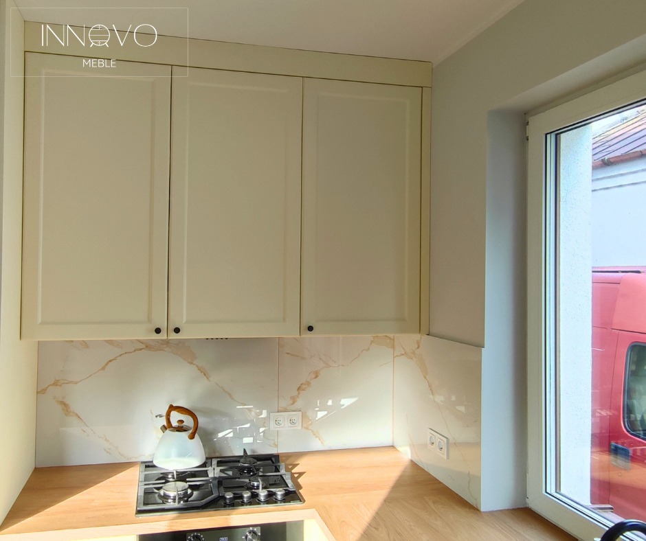 Jasne, kremowe szafki kuchenne z czarnymi uchwytami, marmurowy backsplash i czajnik na płycie gazowej. Widok z okna na zewnątrz.