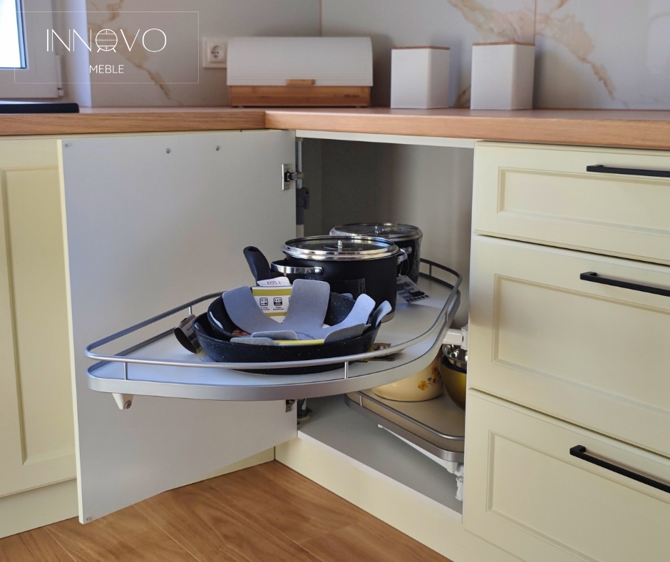 Beżowa kuchnia narożna z wysuwaną półką na garnki i patelnie, czarne uchwyty, drewniany blat i marmurowe płytki w tle. Otwarta szafka narożna.