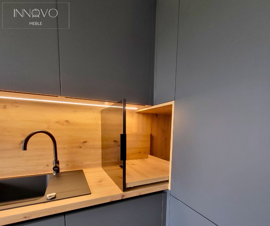 Nowoczesna, szara kuchnia z drewnianym blatem i podświetleniem LED. Umywalka z czarną baterią i otwartą szafką z wysuwaną półką. Minimalistyczny design.
