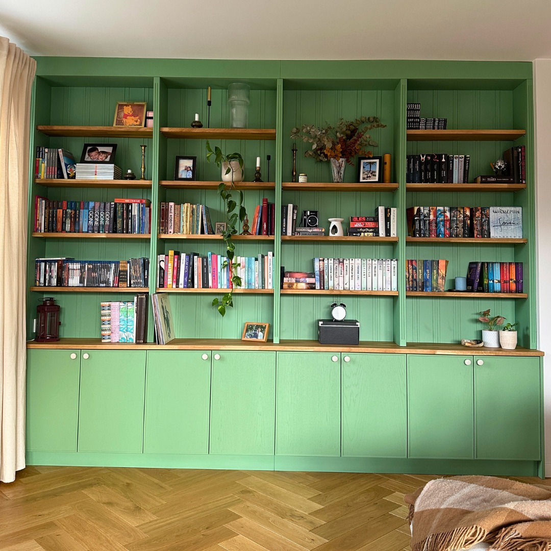 Zielona, zabudowana biblioteczka na wymiar z drewnianymi półkami i szafkami. Ustawione książki, dekoracje i roślina pnąca. Parkiet w jodełkę na podłodze.