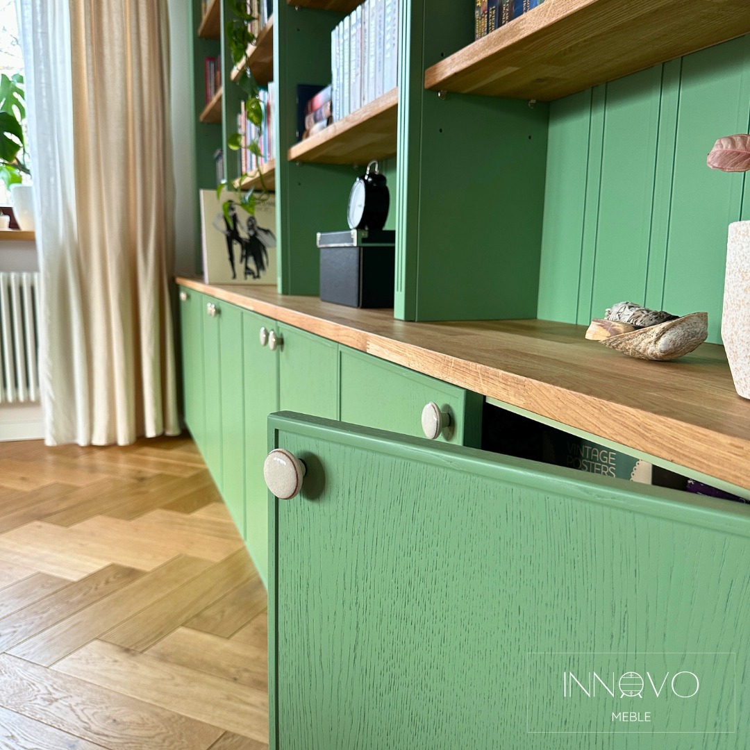 Zielona zabudowa meblowa z drewnianym blatem i otwartą szafką, eksponująca książki. Parkiet w jodełkę i naturalne światło dopełniają wnętrze.
