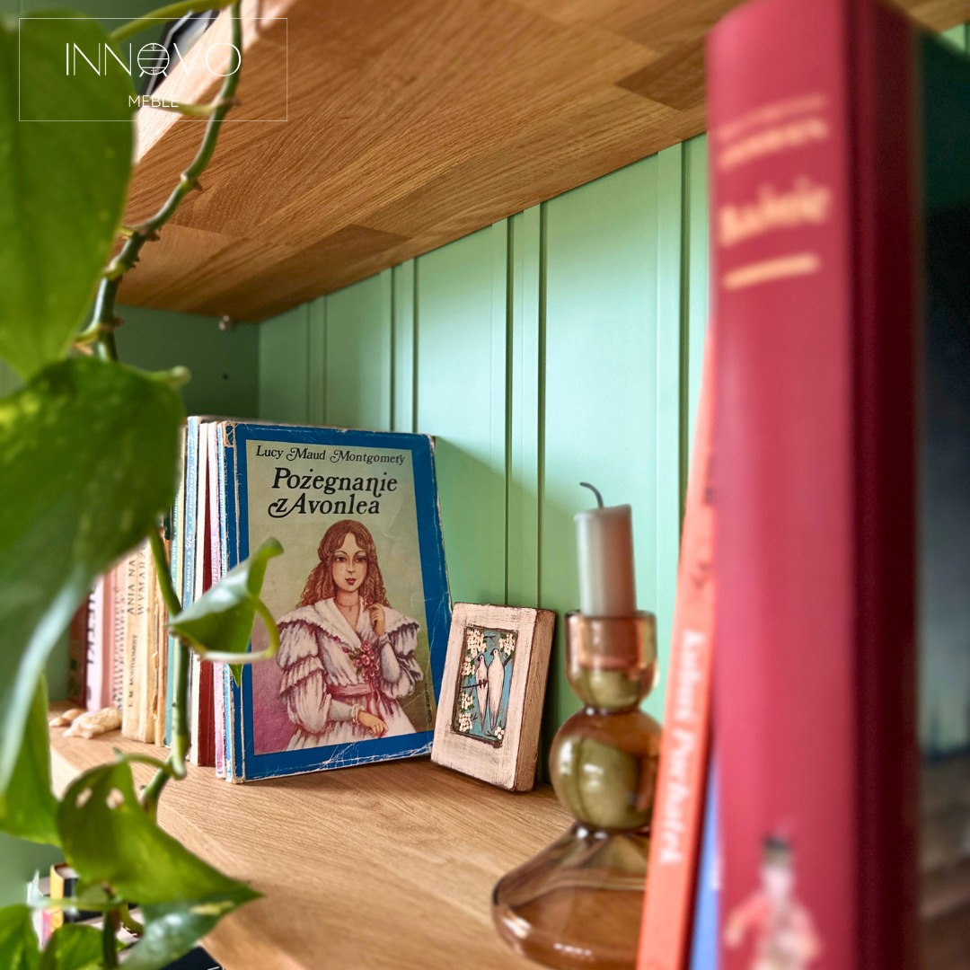 Półka z książkami i dekoracjami na tle zielonej, panelowej ściany. Element rośliny w lewym dolnym rogu kadru dodaje naturalnego akcentu.