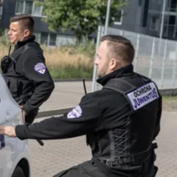 Dwóch ochroniarzy w czarnych uniformach z logo 'Ochrona Juventus' na tle nowoczesnego budynku, jeden stoi z radiem, drugi kuca przy białym samochodzie.