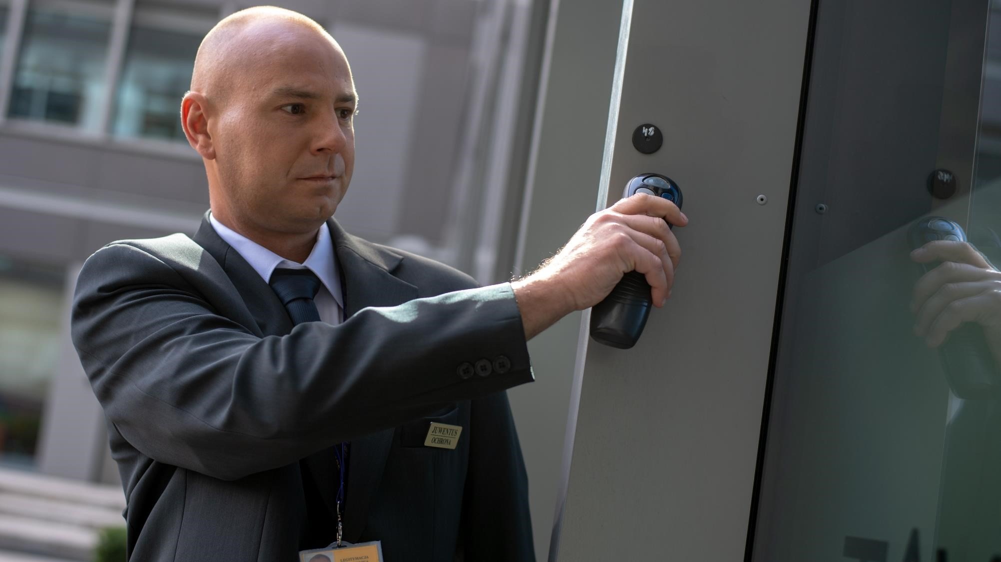 Mężczyzna w garniturze z identyfikatorem 'Juventus Ochrona' używa breloka zbliżeniowego do otwierania przeszklonych drzwi wejściowych do biurowca.