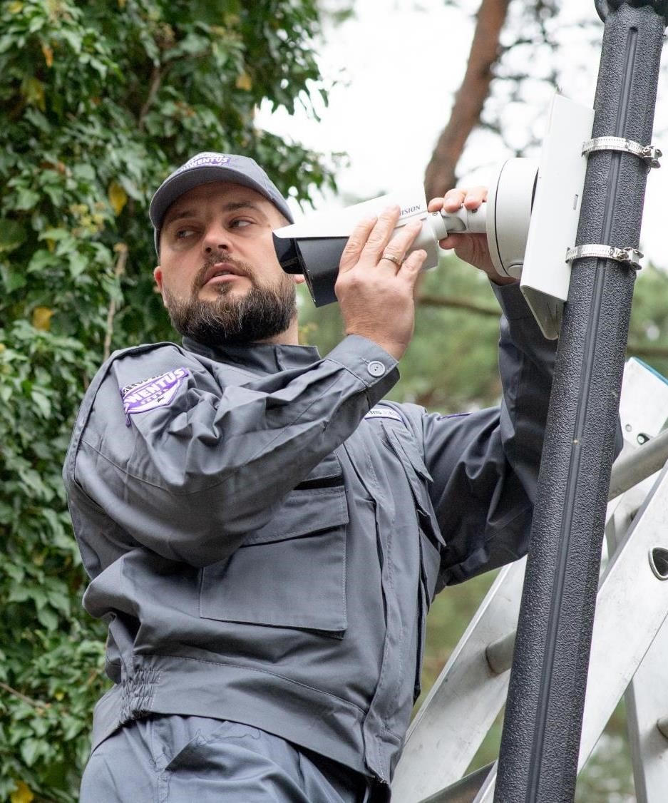 Mężczyzna w szarym uniformie z naszywką firmy VENTUS na drabinie, instaluje kamerę monitoringu na słupie, zabezpieczając ją metalowymi obejmami.