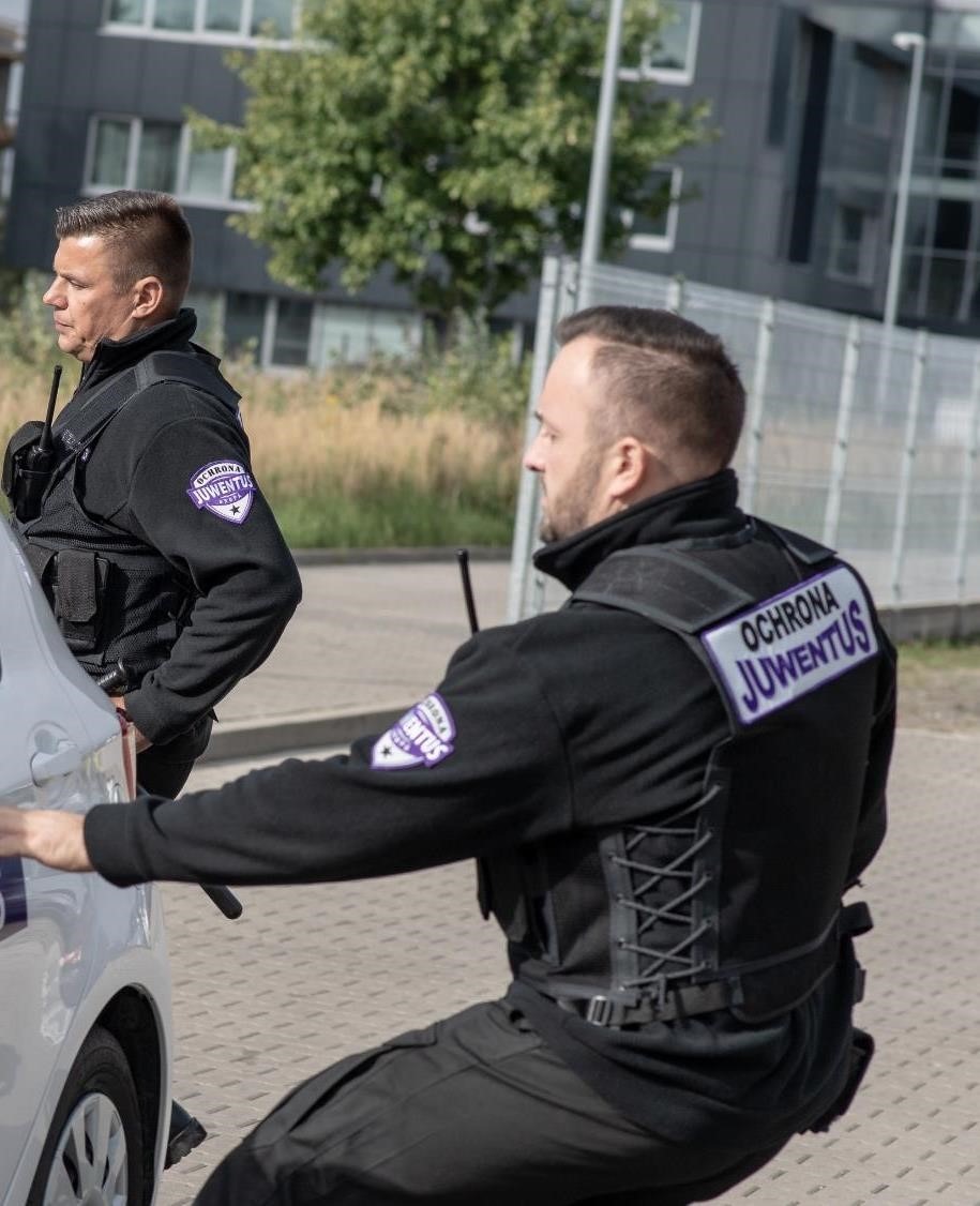 Dwóch ochroniarzy w czarnych uniformach z logo 'Ochrona Juventus' na tle nowoczesnego budynku, jeden stoi z radiem, drugi kuca przy białym samochodzie.