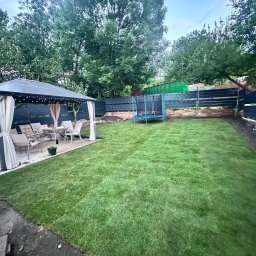 PAWEŁ PASZKOWICZ Rad Nelia - Świeżo założony trawnik z rolki w ogrodzie z altaną i trampoliną. Widoczne ciemne ogrodzenie i drzewa w tle. Częściowo widoczny teren przygotowany pod dalsze prace.