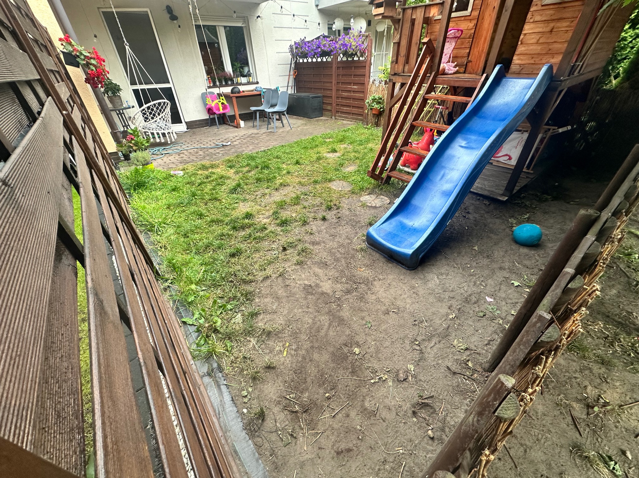 Przygotowanie terenu pod trawnik: wyrównana ziemia z fragmentami trawy, plac zabaw z drewnianą konstrukcją i zjeżdżalnią, widok z perspektywy ogrodzenia.