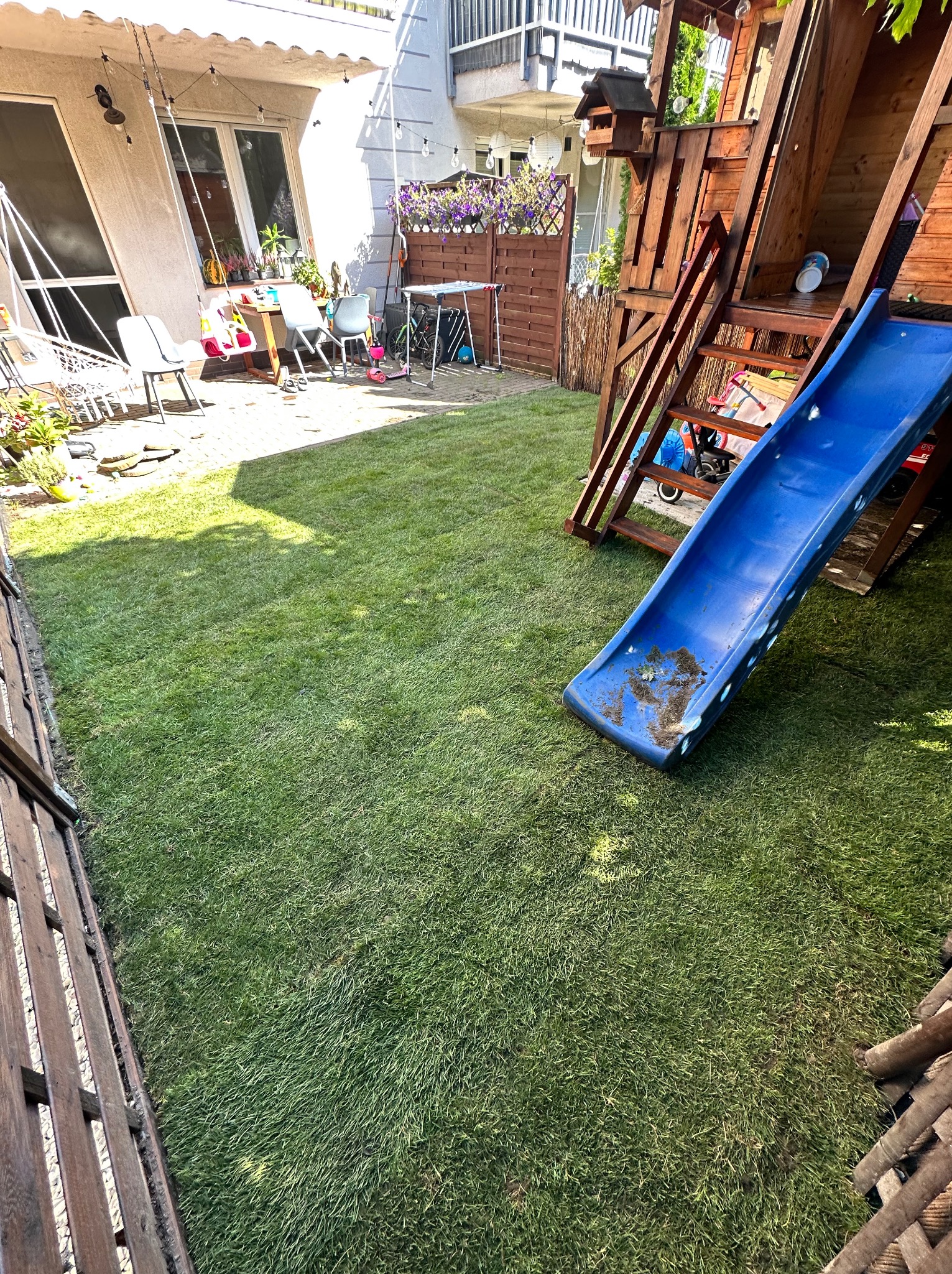 Zadbany trawnik w ogrodzie z drewnianym placem zabaw i zjeżdżalnią. Widoczne meble ogrodowe i dekoracje. Świeżo skoszona trawa, równa powierzchnia.