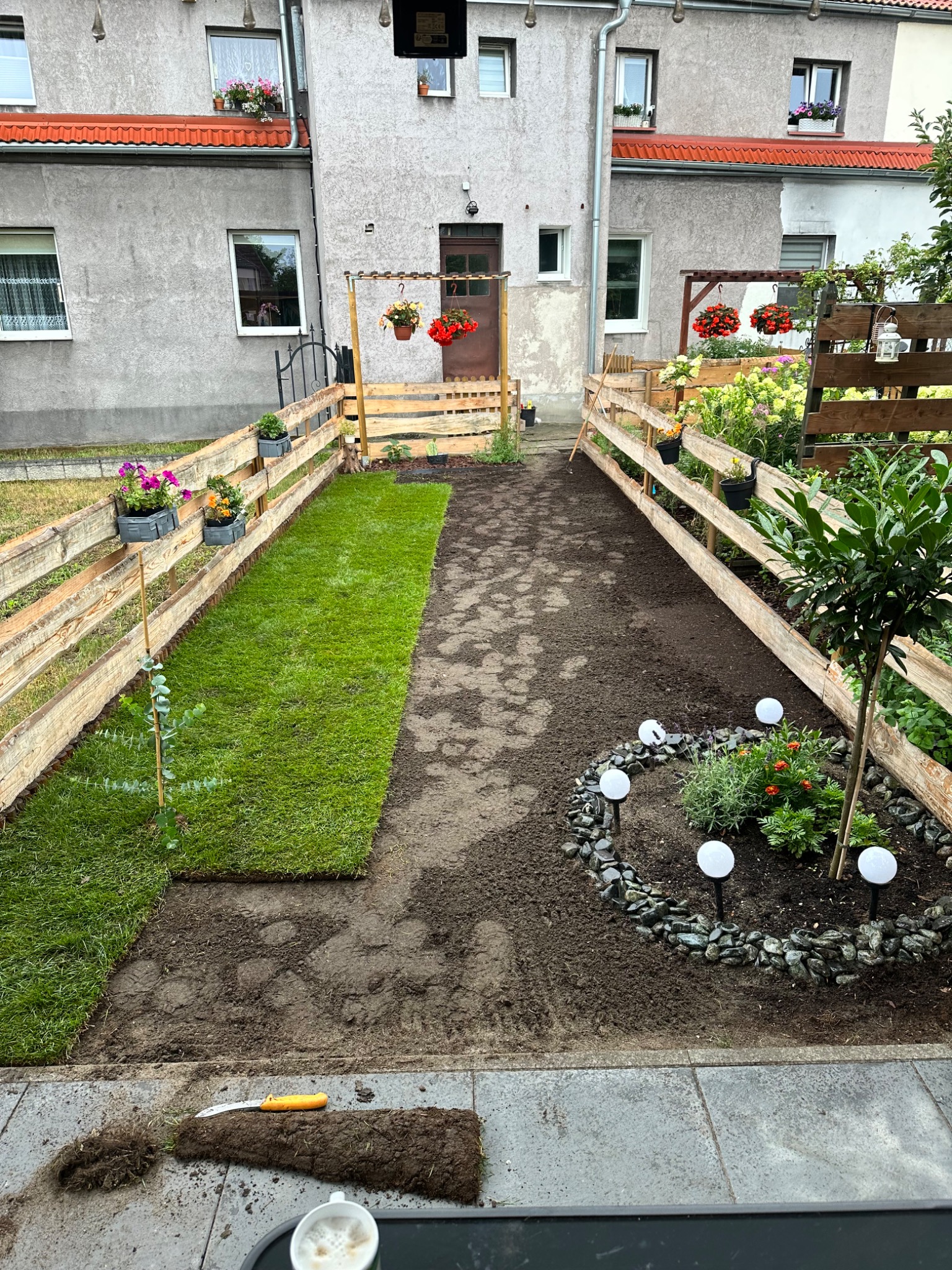 Układanie trawy z rolki w ogrodzie z drewnianym płotem, kwiatami w donicach i dekoracyjnym oświetleniem. Widoczny fragment rolki trawy i narzędzie ogrodnicze.