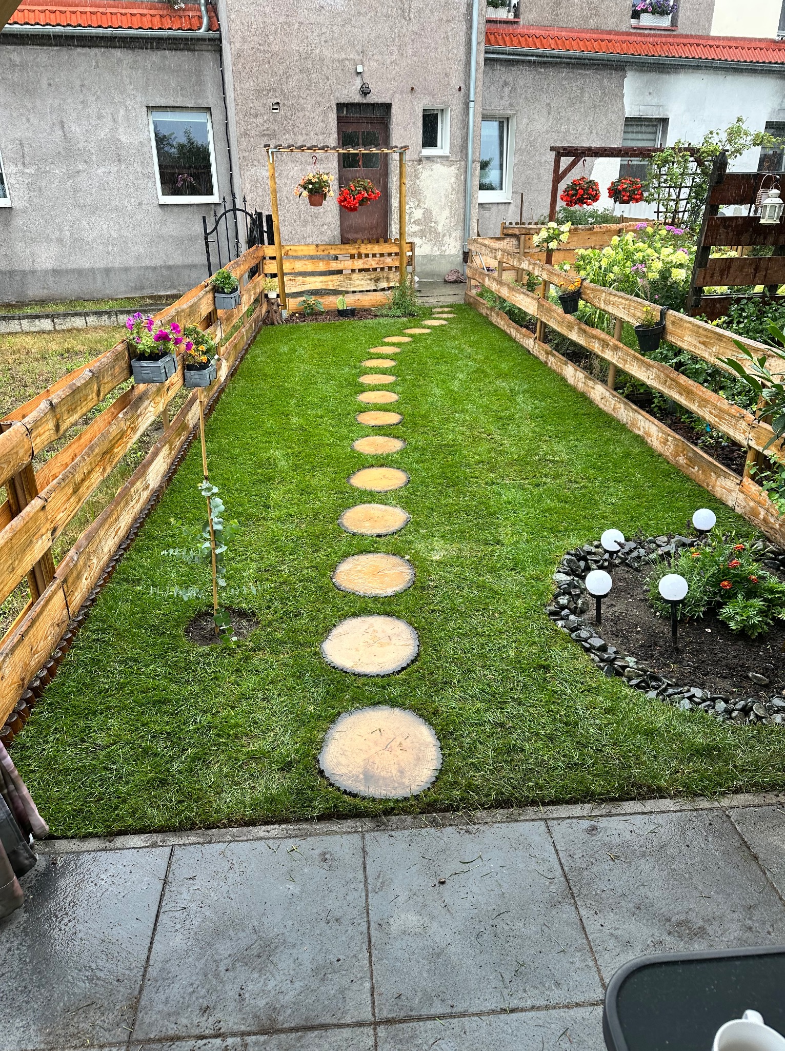 Ułożona trawa z drewnianą ścieżką i ogrodzeniem z palet, dekoracyjne lampy w ogrodzie, kwiaty w donicach. Widok na budynek mieszkalny w tle.