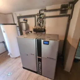 Nowoczesny piec na pellet marki Defro Evopell z zbiornikiem na wodę, zainstalowany w kotłowni z drewnopodobną podłogą i szarymi rurami instalacji grzewczej.