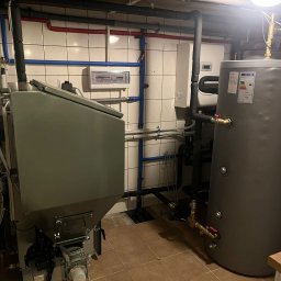 Centrum Ciepła Mariusz Trzęsała - Szary piec na paliwo stałe z zasobnikiem i bojlerem w kotłowni z instalacją grzewczą na ścianie z płytek. Widoczne rury w kolorze niebieskim i czerwonym.