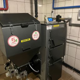 Centrum Ciepła Mariusz Trzęsała - Nowoczesny piec na paliwo stałe klasy 5 z podajnikiem, oznaczony naklejkami 'eco design' i 'suchy opał', zainstalowany w kotłowni z wykafelkowanymi ścianami.