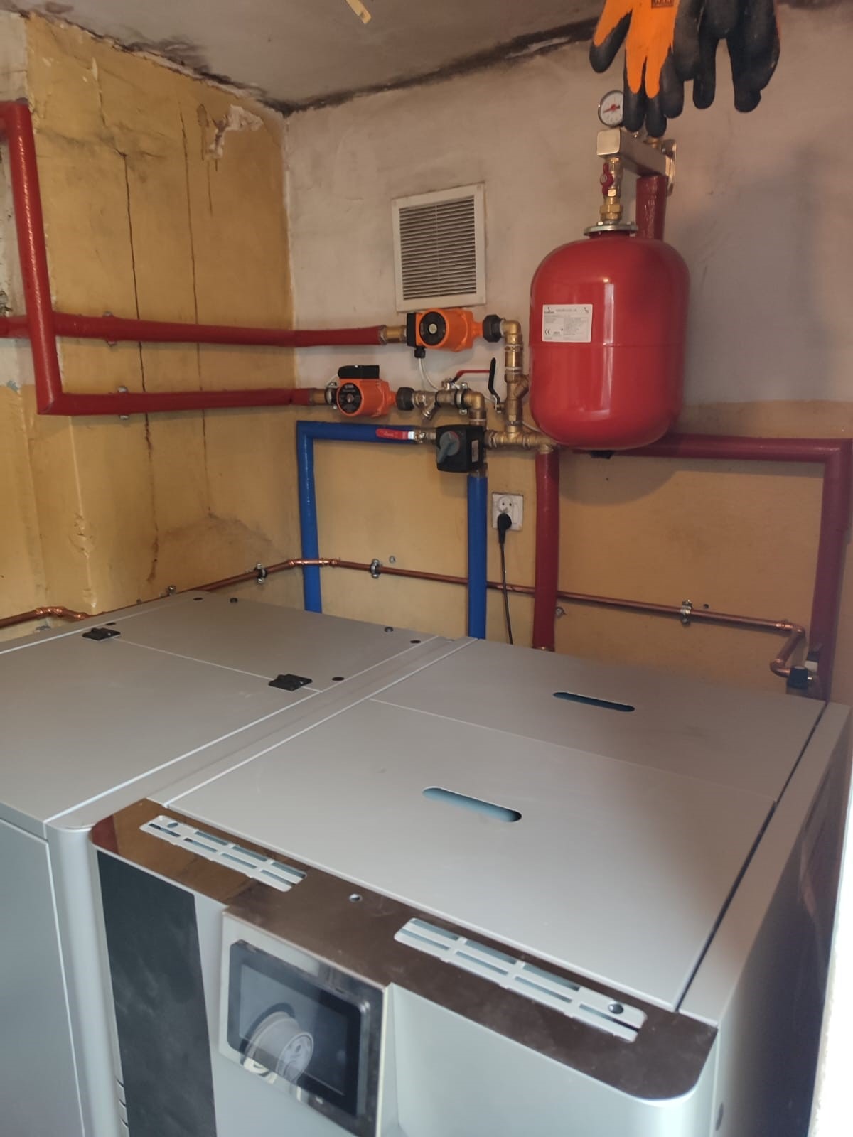 Szary piec grzewczy z podłączoną instalacją: czerwone rury, pompy obiegowe i naczynie wzbiorcze, rękawice robocze wiszące na rurach.