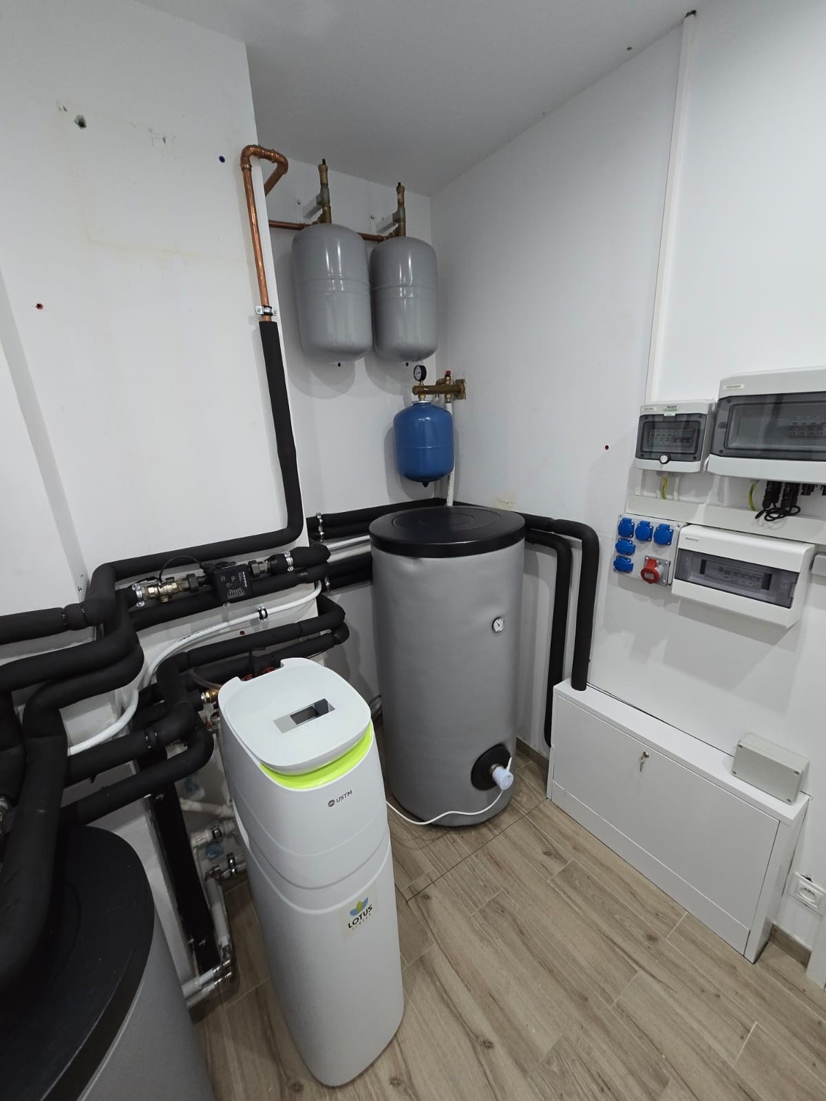 Szara instalacja grzewcza z zbiornikiem akumulacyjnym, zmiękczaczem wody Lotus i rozdzielnicą elektryczną w pomieszczeniu z panelami podłogowymi imitującymi drewno.
