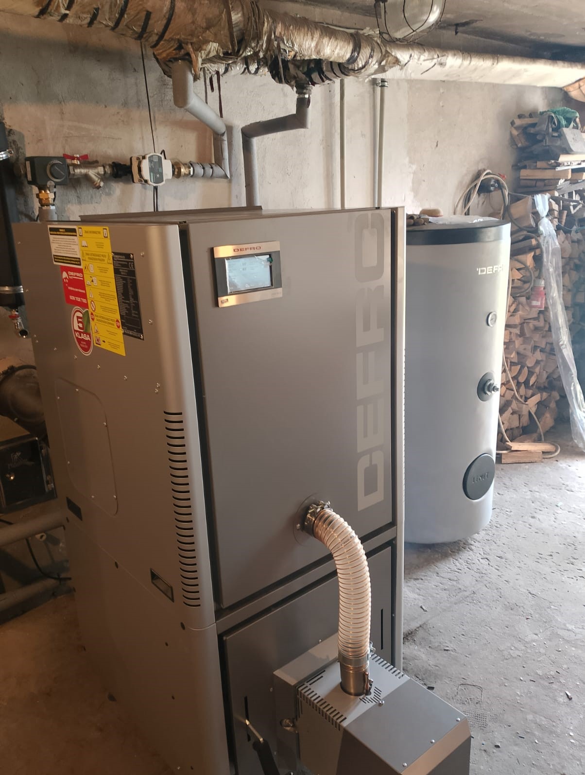 Szary piec na paliwo stałe Defro z podajnikiem i zasobnikiem na paliwo, widoczny także zasobnik ciepłej wody użytkowej, instalacja w kotłowni.