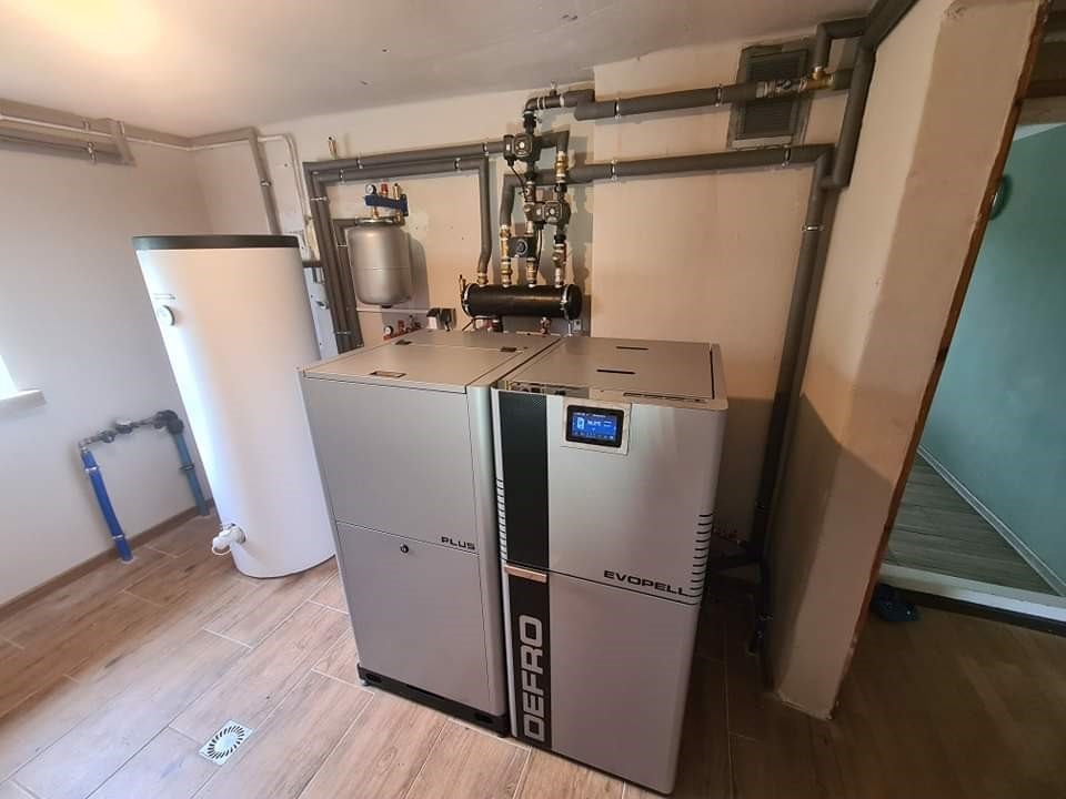 Nowoczesny piec na pellet marki Defro Evopell z zbiornikiem na wodę, zainstalowany w kotłowni z drewnopodobną podłogą i szarymi rurami instalacji grzewczej.