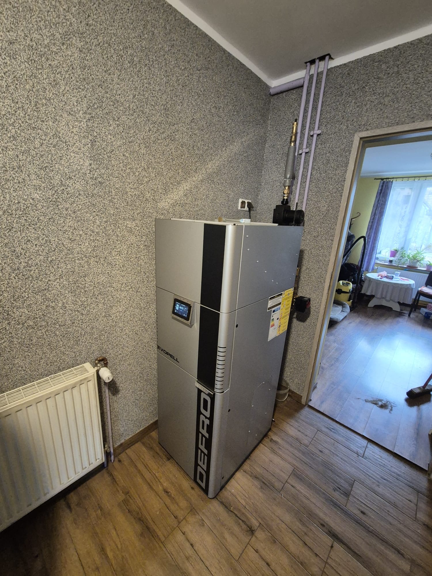 Szary piec Defro Evopell w rogu pokoju z grzejnikiem i drewnopodobną podłogą. Widoczne rury instalacji grzewczej i fragment otwartego przejścia do innego pomieszczenia.