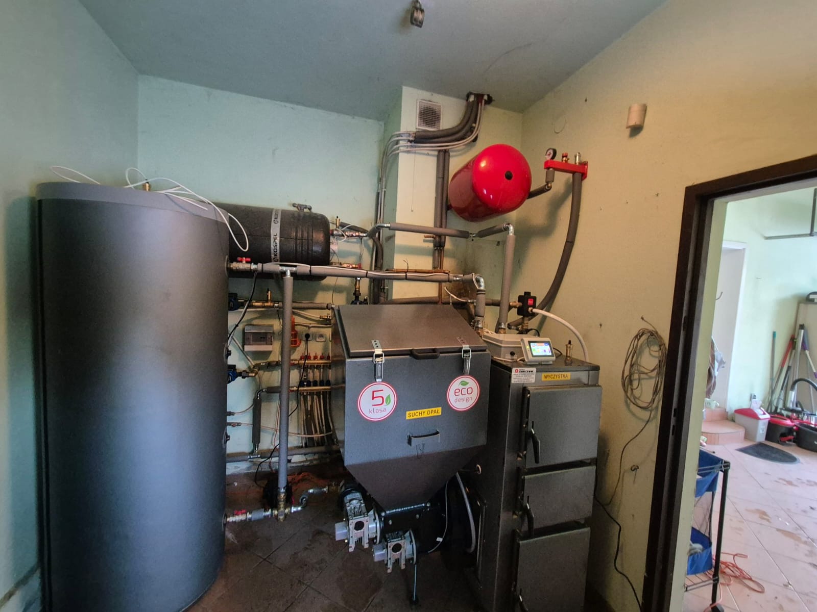 Zmontowany, nowoczesny piec na paliwo stałe z zasobnikiem, widoczne rury, zbiornik wyrównawczy i sterownik. Całość w kotłowni o jasnych ścianach.