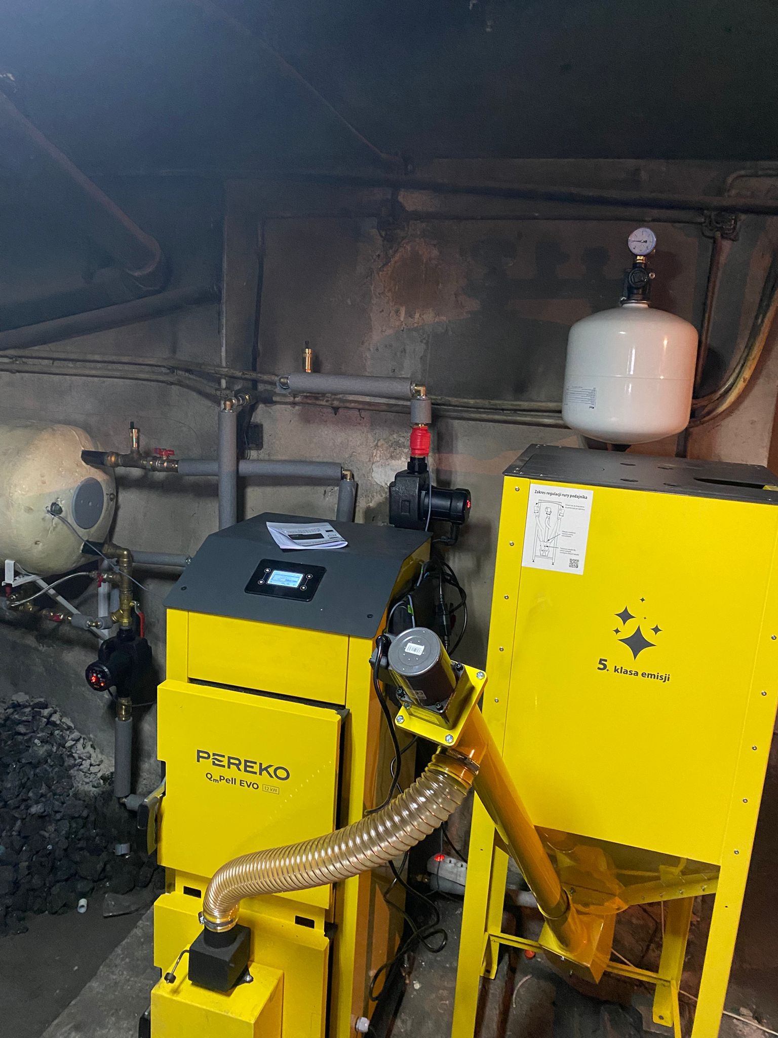 Żółty piec Pereko QmPell EVO 12 kW z podajnikiem, widoczne rury instalacji CO i zbiornik wyrównawczy w kotłowni. 5 klasa emisji.