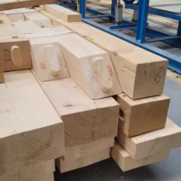 Stos jasnej tarcicy budowlanej z widocznymi oznaczeniami 'HOUSE', '609' i '612' naniesionymi czerwoną farbą, ułożony w hali produkcyjnej z niebieską konstrukcją w tle.