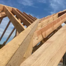 Konstrukcja drewniana dachu w trakcie montażu, widok z dołu na krokwie i łaty na tle błękitnego nieba.