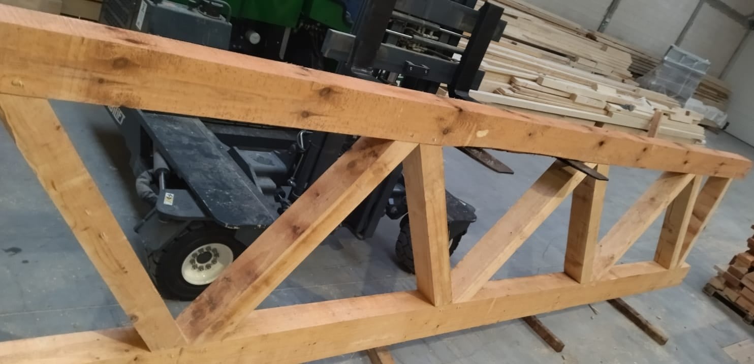 Gotowa drewniana konstrukcja kratowa więźby dachowej, leżąca na metalowych wspornikach w hali magazynowej, w tle wózek widłowy i stosy tarcicy.