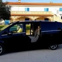 Czarny, lśniący Mercedes-Benz Vito z otwartymi drzwiami przesuwnymi, zaparkowany na żwirowym podjeździe przed dwupiętrowym budynkiem z białymi ścianami i niebieskimi okiennicami, otoczony zielenią.