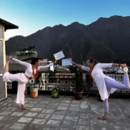 Dwie kobiety w białych strojach i pomarańczowych girlandach wykonują pozycję taneczną na dachu z widokiem na góry, trzymając certyfikaty.