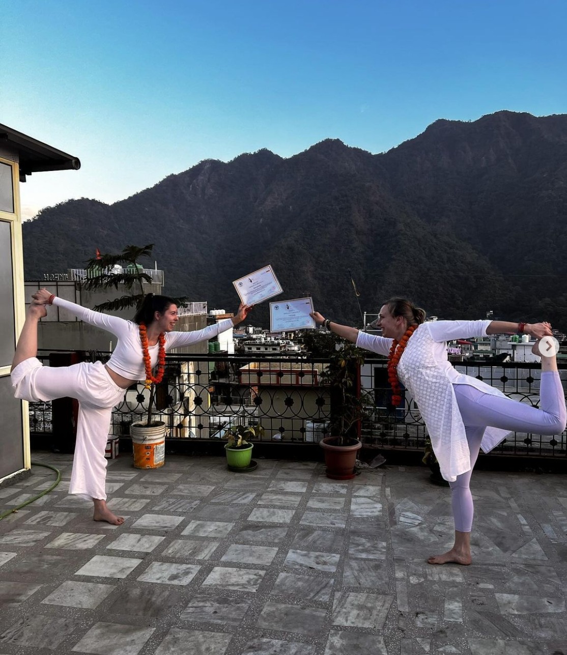 Dwie kobiety w białych strojach i pomarańczowych girlandach wykonują pozycję taneczną na dachu z widokiem na góry, trzymając certyfikaty.