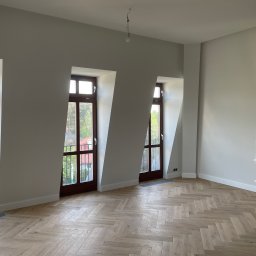 VASBUD - Pusty pokój po remoncie z parkietem ułożonym we wzór jodełki francuskiej, trzema oknami balkonowymi z ciemnego drewna i świeżo pomalowanymi ścianami na szaro. W rogu widoczny sprzęt AGD...