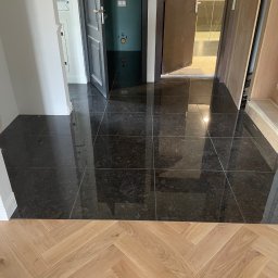 VASBUD - Połączenie ciemnej, błyszczącej posadzki z płytek i jasnego parkietu ułożonego w jodełkę, widoczne otwarte drzwi i fragmenty ścian w trakcie wykańczania.