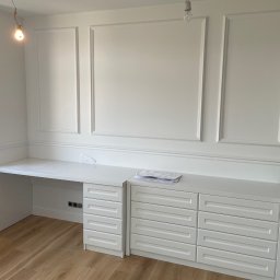 VASBUD - Białe, zabudowane biurko z szufladami, wkomponowane w ścianę z dekoracyjnymi listwami, na podłodze panele, surowa żarówka zwisa z sufitu.