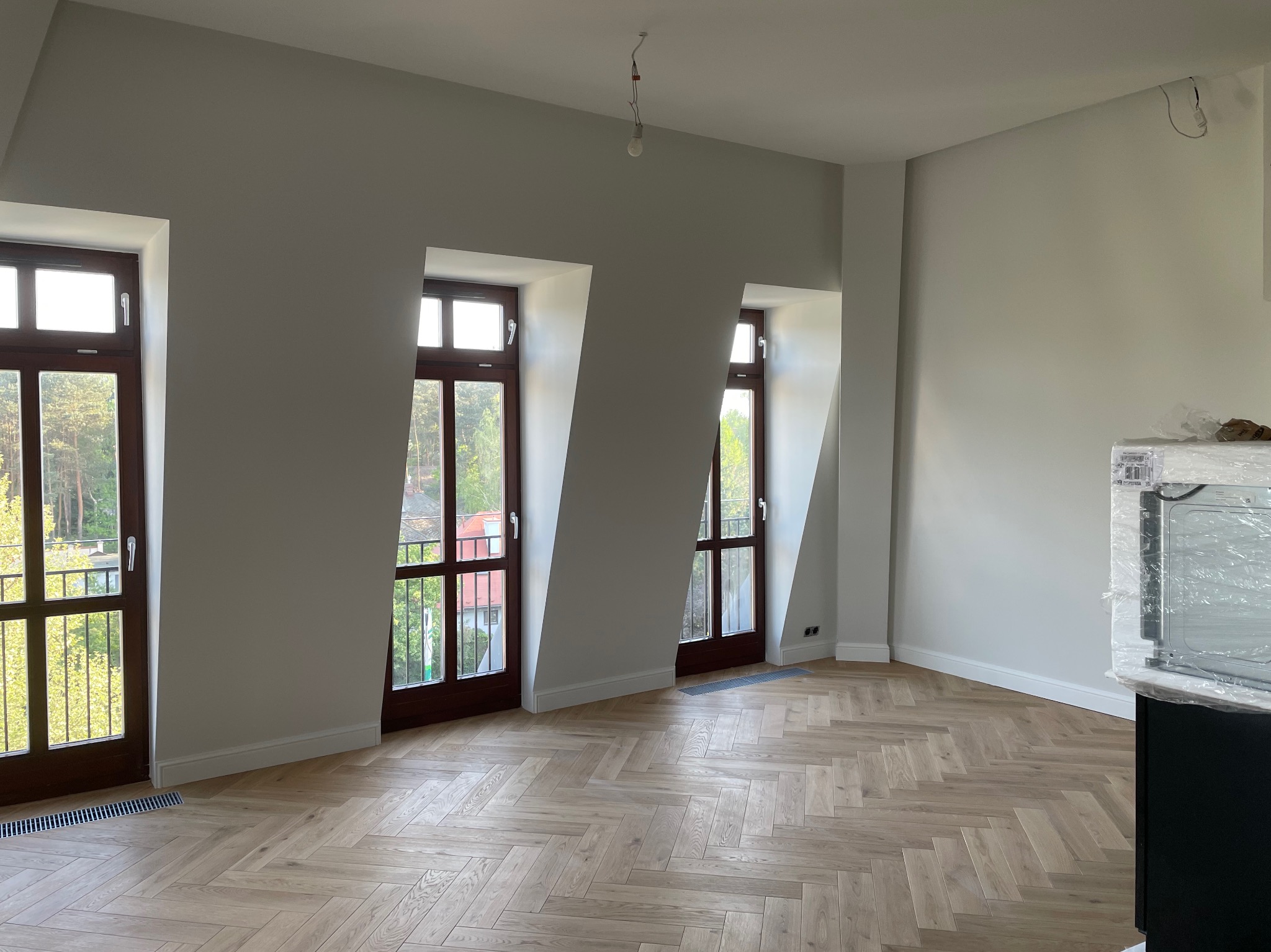 Pusty pokój po remoncie z parkietem ułożonym we wzór jodełki francuskiej, trzema oknami balkonowymi z ciemnego drewna i świeżo pomalowanymi ścianami na szaro. W rogu widoczny sprzęt AGD...