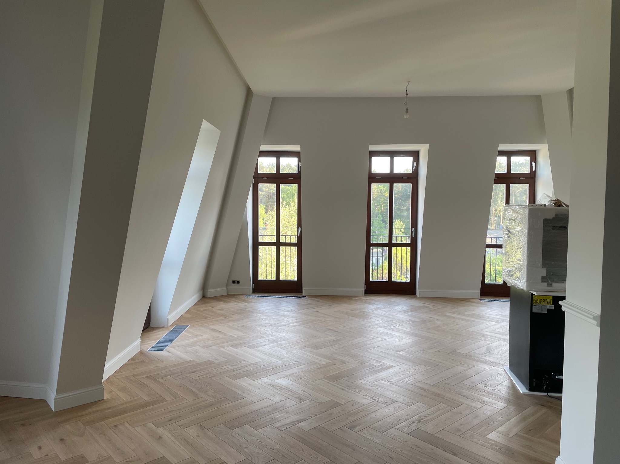 Jasne, przestronne poddasze po remoncie, z parkietem ułożonym w jodełkę, trzema drewnianymi oknami balkonowymi i świeżo pomalowanymi ścianami na neutralny odcień szarości.