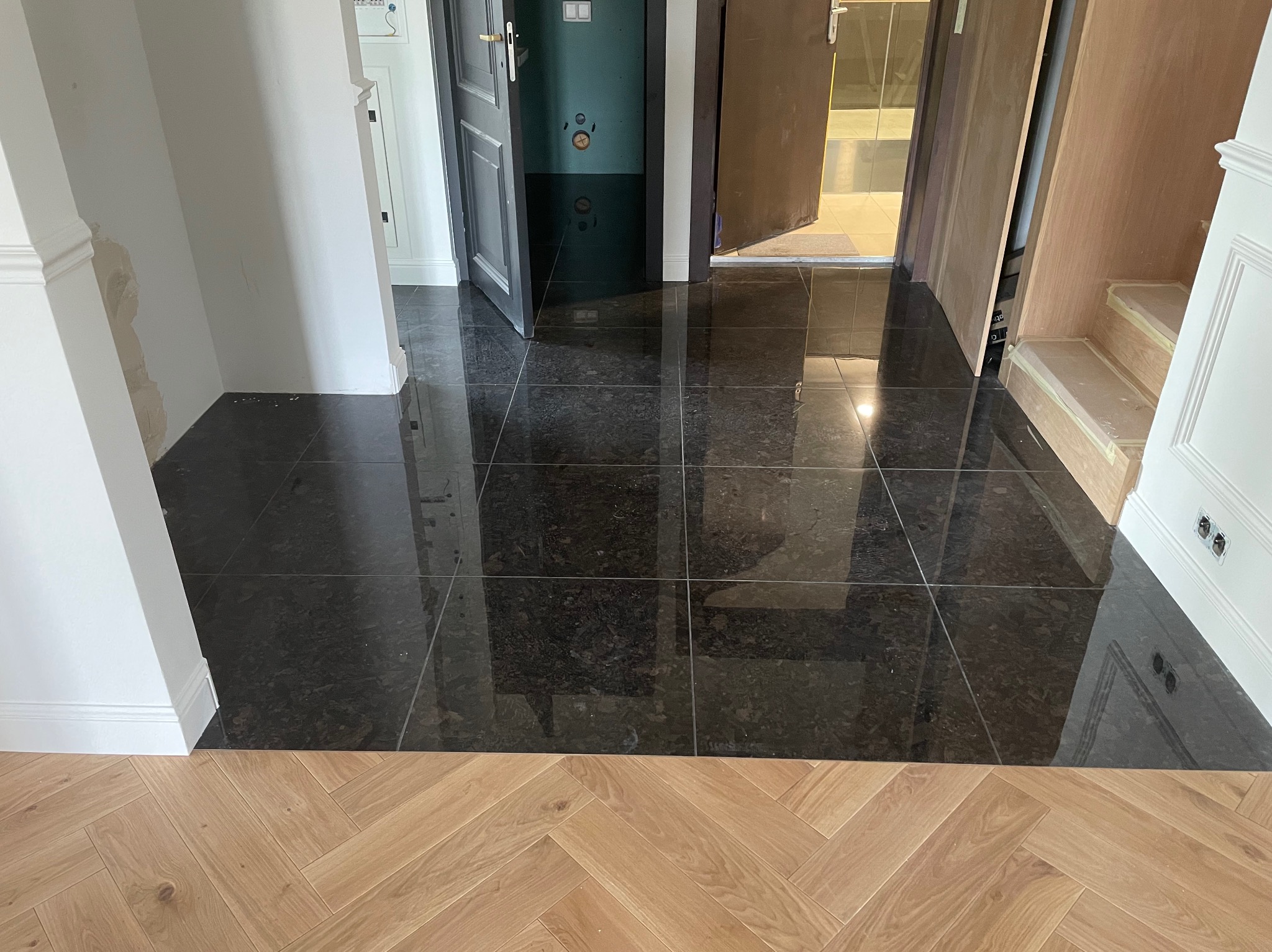 Połączenie ciemnej, błyszczącej posadzki z płytek i jasnego parkietu ułożonego w jodełkę, widoczne otwarte drzwi i fragmenty ścian w trakcie wykańczania.