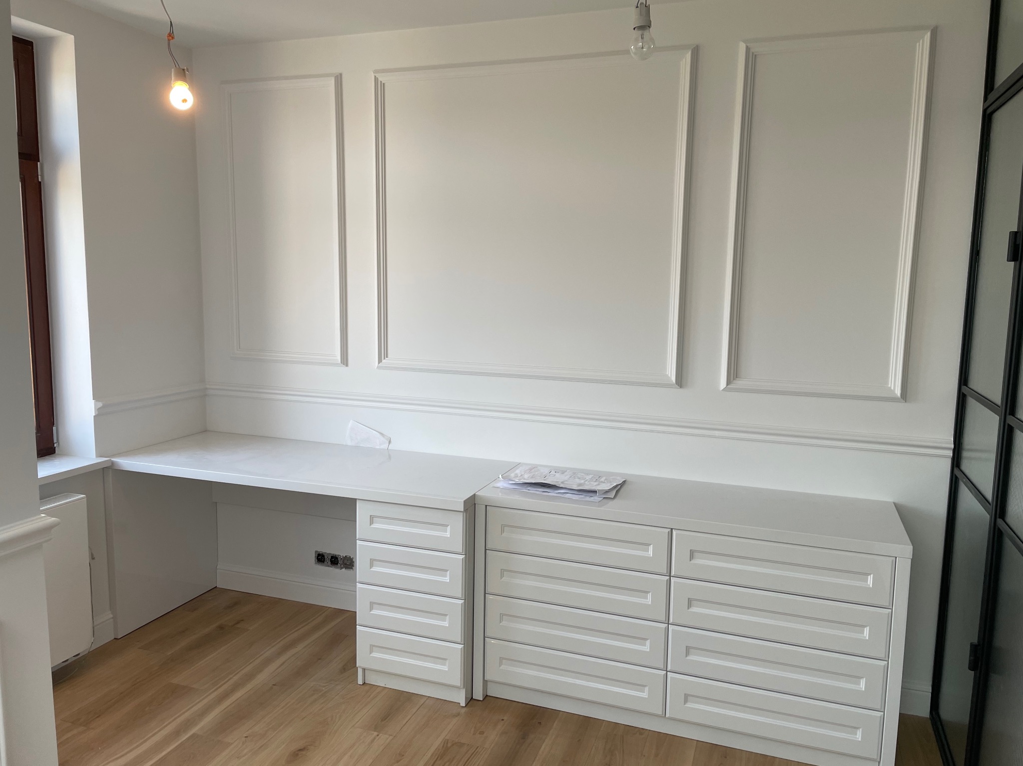 Białe, zabudowane biurko z szufladami, wkomponowane w ścianę z dekoracyjnymi listwami, na podłodze panele, surowa żarówka zwisa z sufitu.