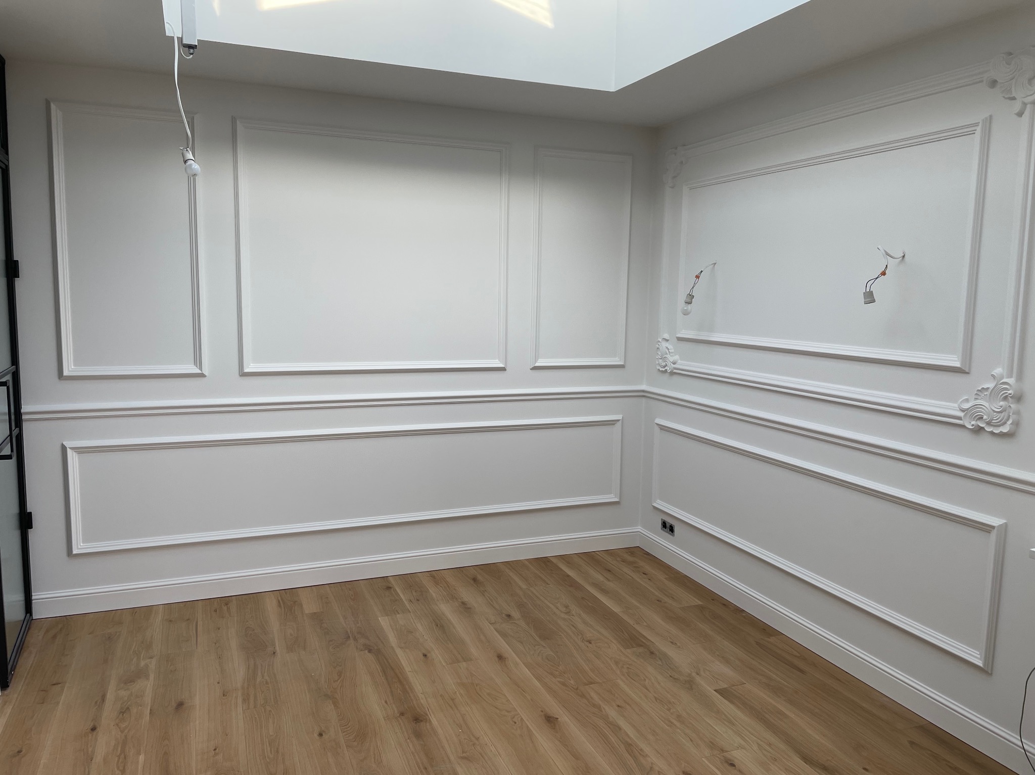 Białe ściany z dekoracyjnymi sztukateriami i drewnianą podłogą, widoczne kable elektryczne wystające ze ściany, surowe wykończenie w nowym pomieszczeniu z oknem dachowym.