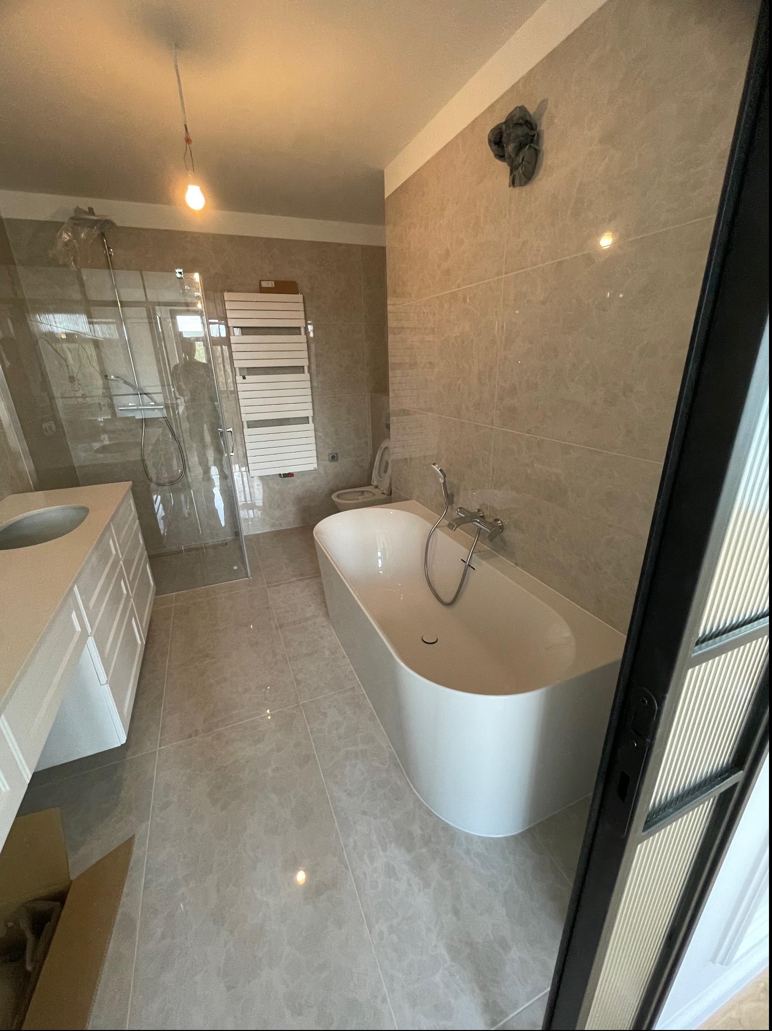 Nowoczesna łazienka z wanną wolnostojącą, kabiną prysznicową ze szklaną ścianką, umywalką z szafką w zabudowie i grzejnikiem drabinkowym, wykończona płytkami w odcieniach szarości; widoczny...