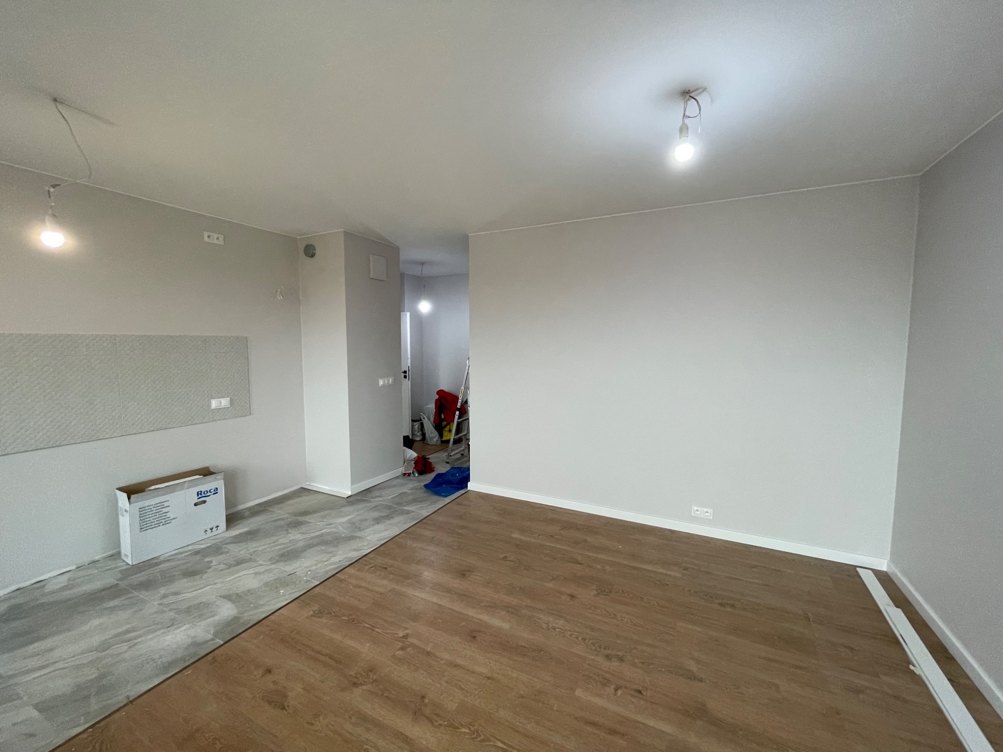Fragment pokoju w trakcie remontu: ułożone szare płytki podłogowe przechodzą w drewnopodobny panel, widoczna kartonowa paczka po płytkach Roca przy ścianie, surowa żarówka na suficie.